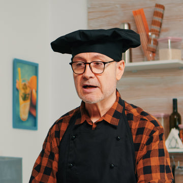 Man in a kitchen wearing a black apron and glasses, holding a metal bowl.